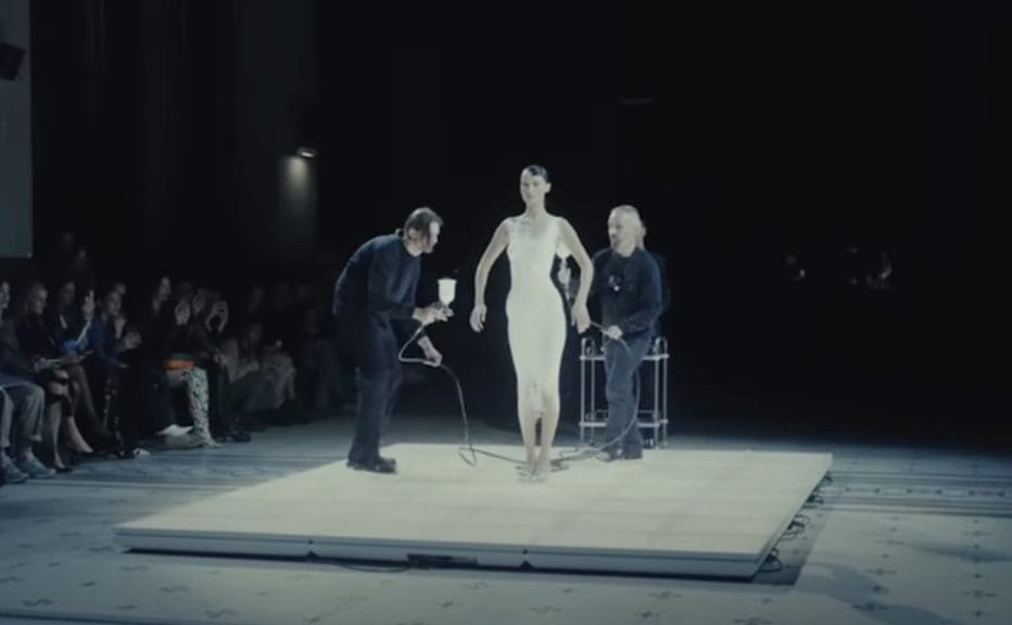 Bella Hadid at the Coperni S/S '23 Paris Fashion Week show having a dress sprayed onto her by two men. A white liquid fibre is sprayed onto her as she's standing still. 